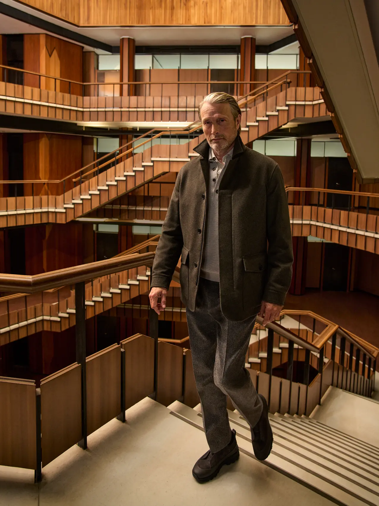 Mads Mikkelsen wearing a dark olive field jacket layered over a knit with tailored trousers and Triple Stitch shoes, on the staircase of a wood-paneled interior atrium, from the ZEGNA Fall/Winter 2026 collection.