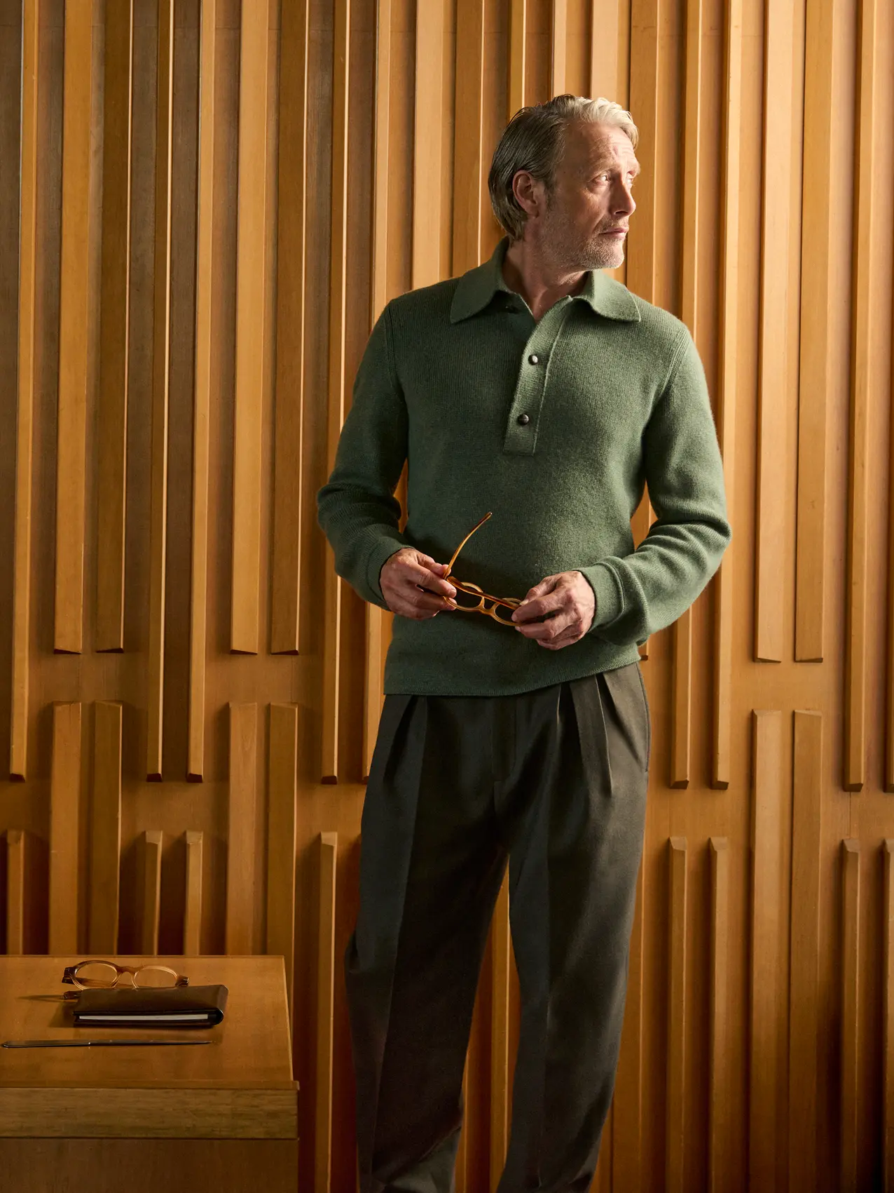 Mads Mikkelsen wearing a green ribbed knit polo with dark tailored trousers, holding his glasses in an elegant wood-paneled interior, from the ZEGNA Fall/Winter 2026 collection.