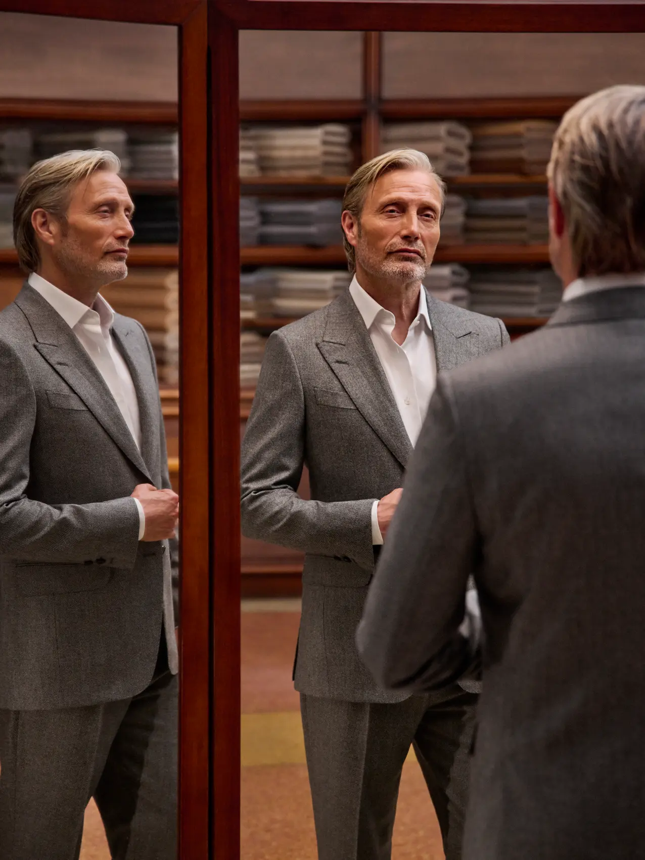 Mads Mikkelsen in a refined grey tailored suit with a white shirt, seen in full in the triple mirror of a tailoring atelier, from the ZEGNA Fall/Winter 2026 collection.