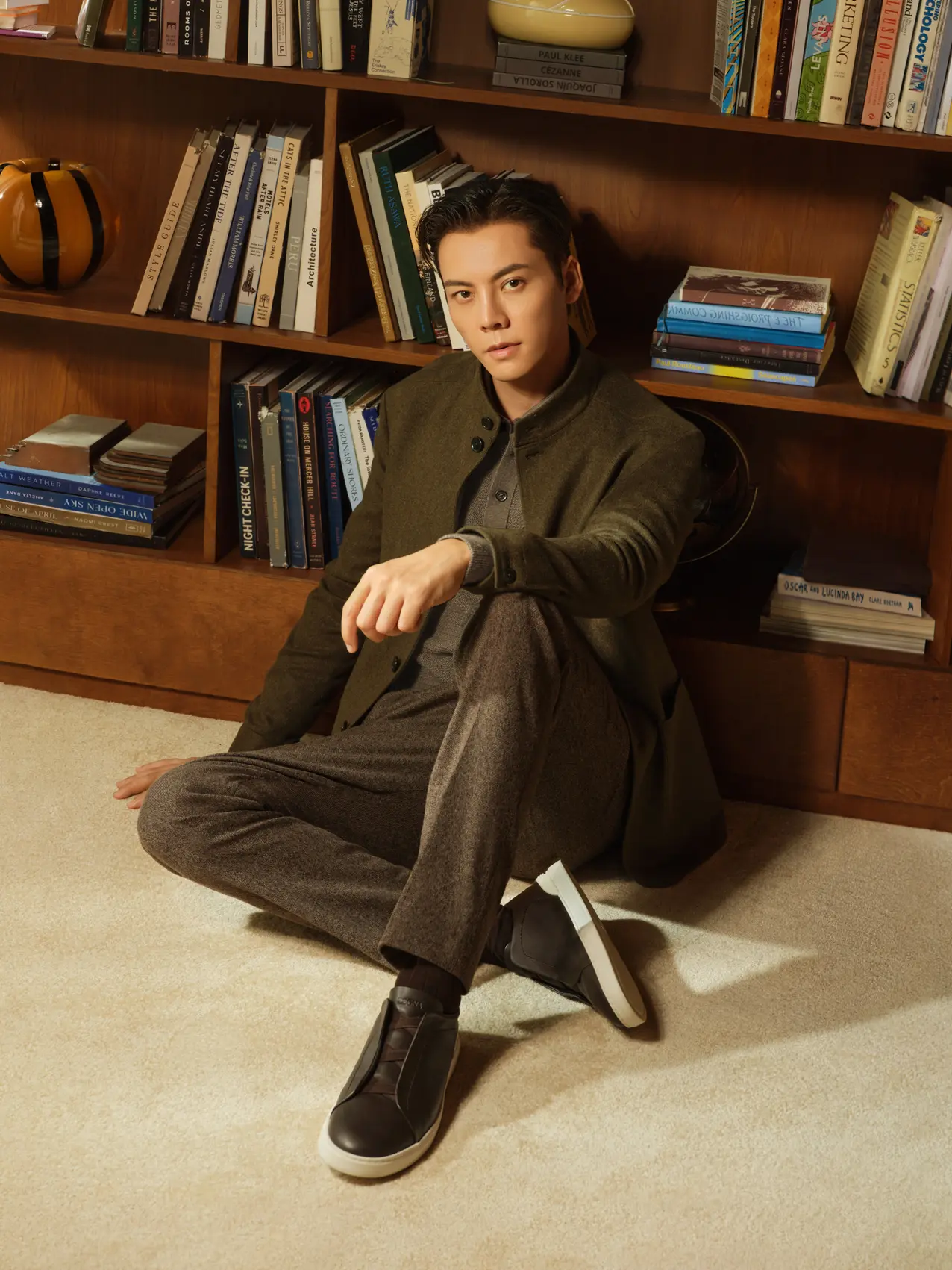 William Chan seated on the floor against a bookshelf, in a refined winter look by Zegna with the Il Conte jacket in Oasi Cashmere, dark olive green multimélange, FW26.