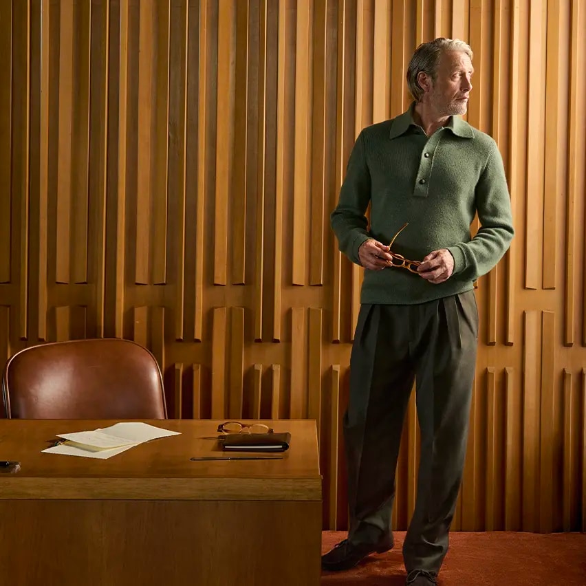 Mads Mikkelsen wearing a green ribbed knit polo with dark tailored trousers, holding his glasses in an elegant wood-paneled interior, from the ZEGNA Fall/Winter 2026 collection.