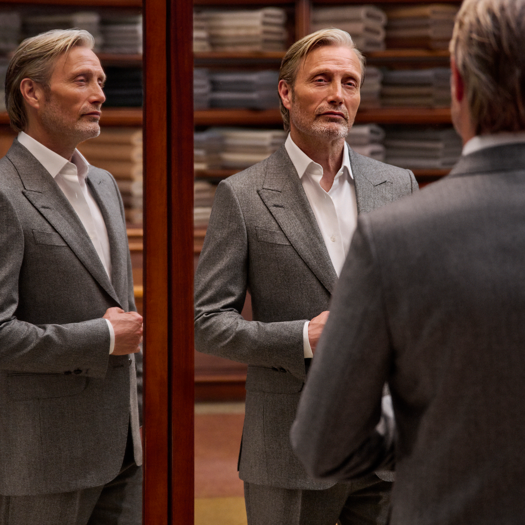 Mads Mikkelsen 身穿灰色精裁西裝配白色恤衫，在裁縫工坊的三面鏡中映出全身造型，展現 ZEGNA 2026 秋冬系列之作。