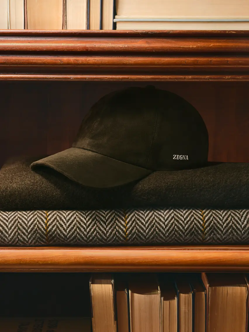ZEGNA（杰尼亚）棒球帽置于木质书架上叠放的围巾之上，来自 ZEGNA（杰尼亚）2026 秋冬系列。