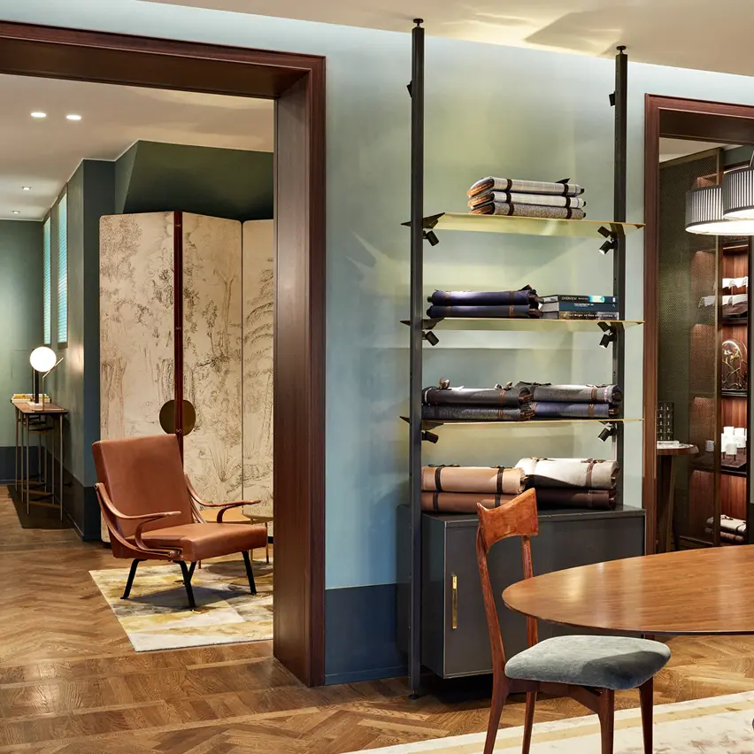 Luxury men's boutique with a white carpet and leather armchairs, showcasing folded clothes on open shelves and a cozy seating area with a wooden table and chairs.