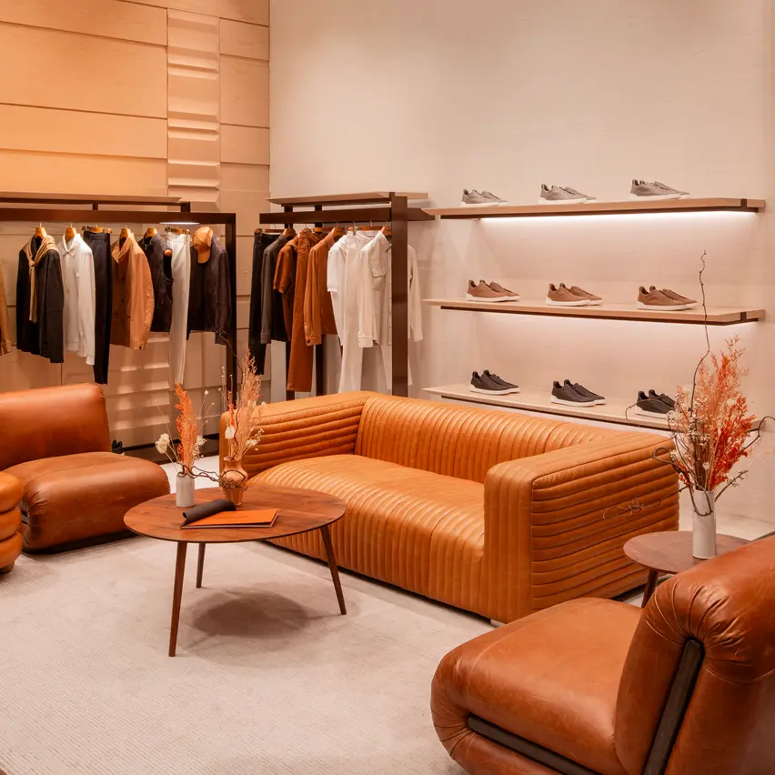 Luxury men's boutique with a white carpet and leather armchairs. Shelves display ZEGNA sneakers, while jackets and coats are elegantly hung on a stand.