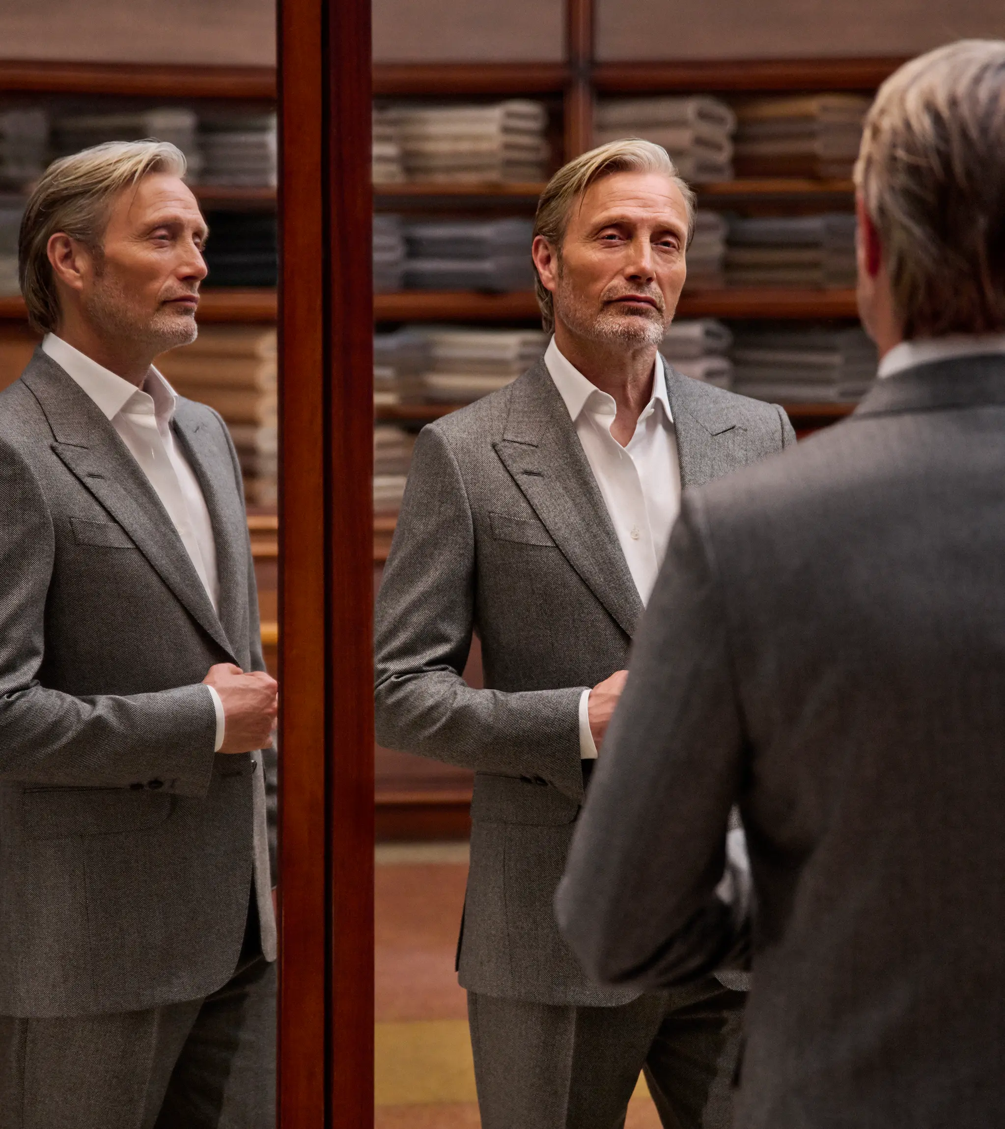 Mads Mikkelsen in a refined grey tailored suit with a white shirt, seen in full in the triple mirror of a tailoring atelier, from the ZEGNA Fall/Winter 2026 collection.