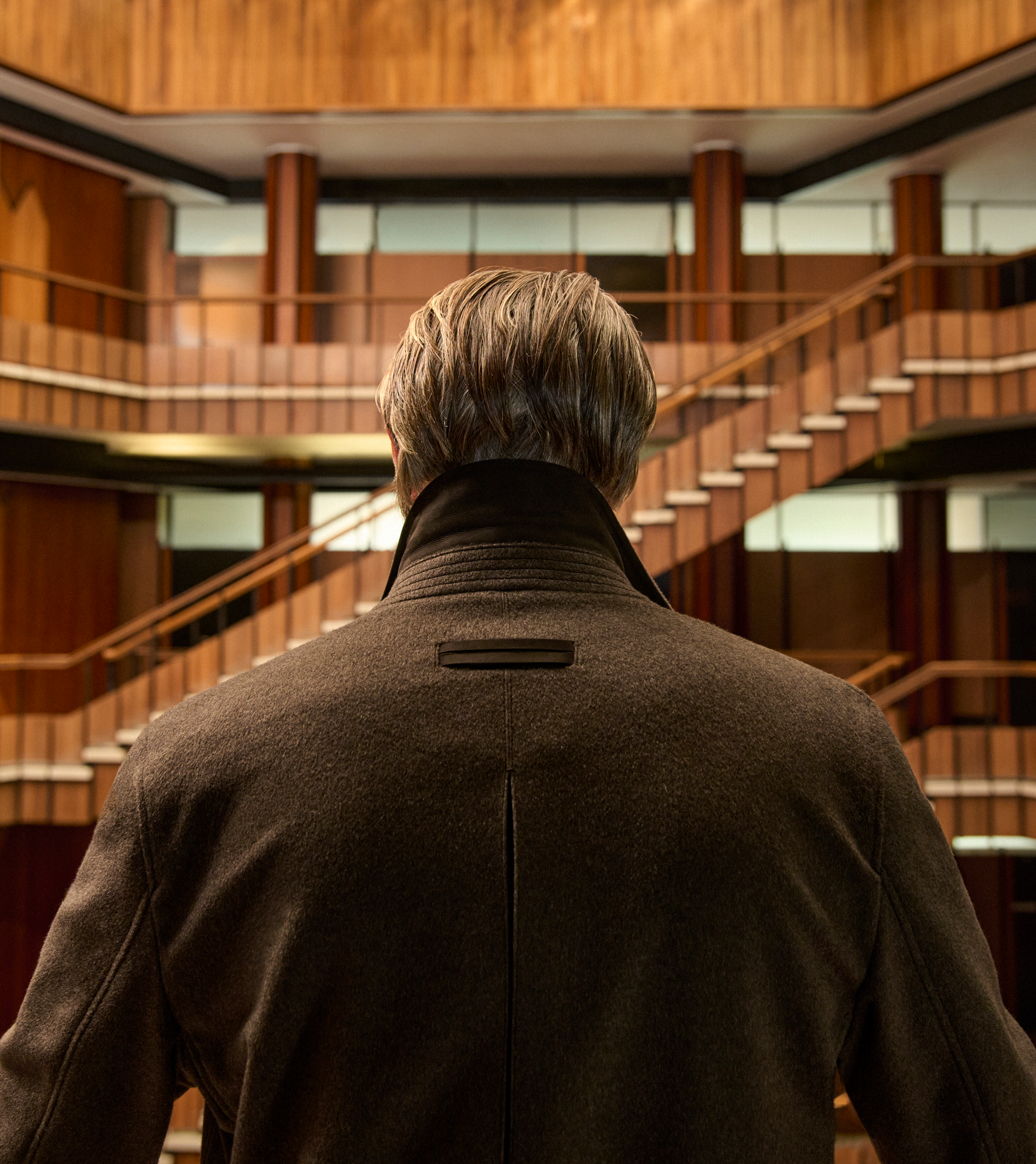 Mads Mikkelsen 身穿深啡羊毛大衣的背影，站在木鑲板室內中庭，展現 ZEGNA 2026 秋冬系列的魅力風采。
