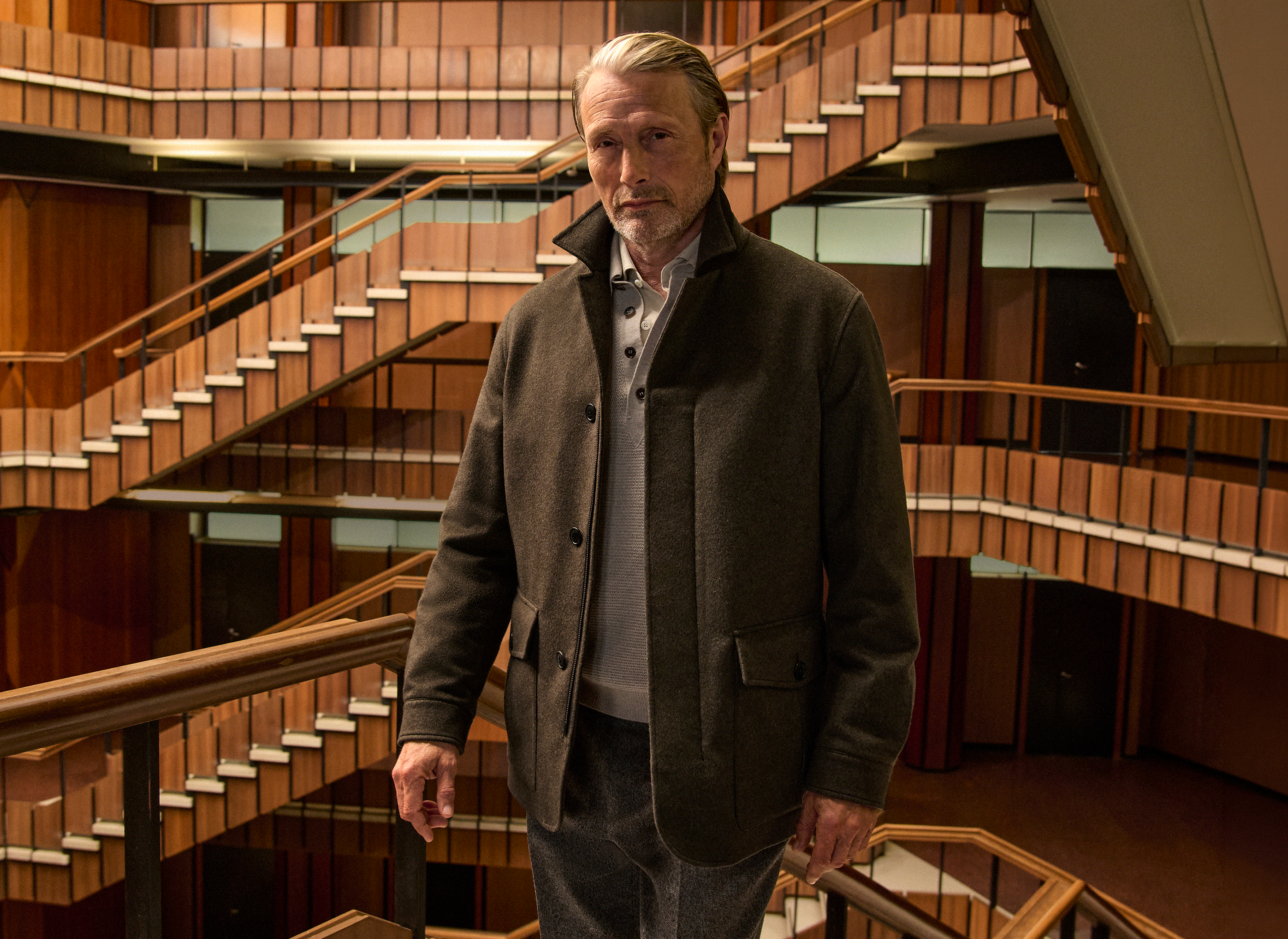Mads Mikkelsen 穿上深橄欖綠色軍䄛，內疊針織衫，配襯剪裁長褲及 Triple Stitch 鞋履，立於木鑲板室內中庭的樓梯上，出自 ZEGNA 2026 秋冬系列。