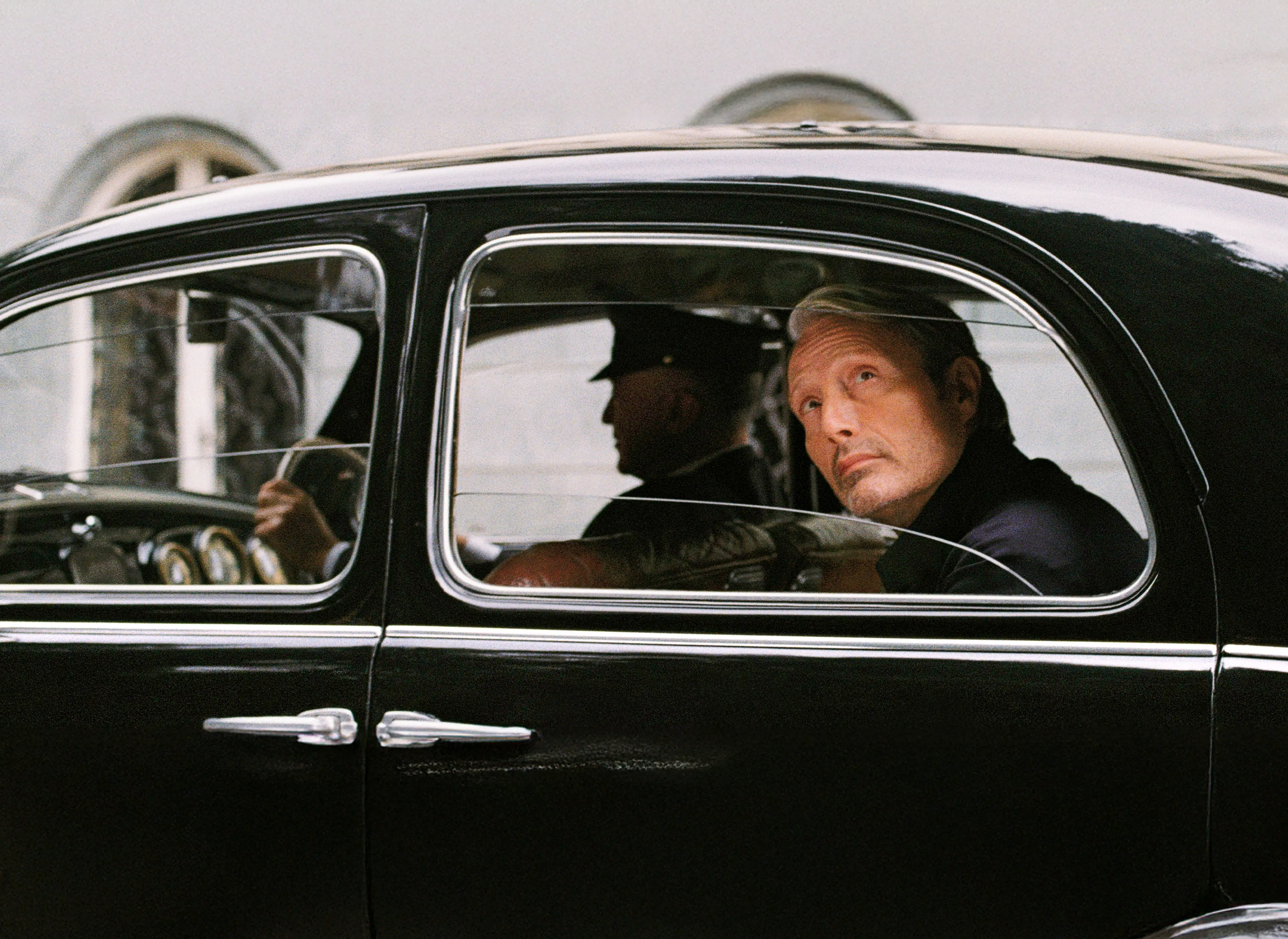 Mads Mikkelsen 坐在經典黑色汽車中，正從側窗凝視窗外。