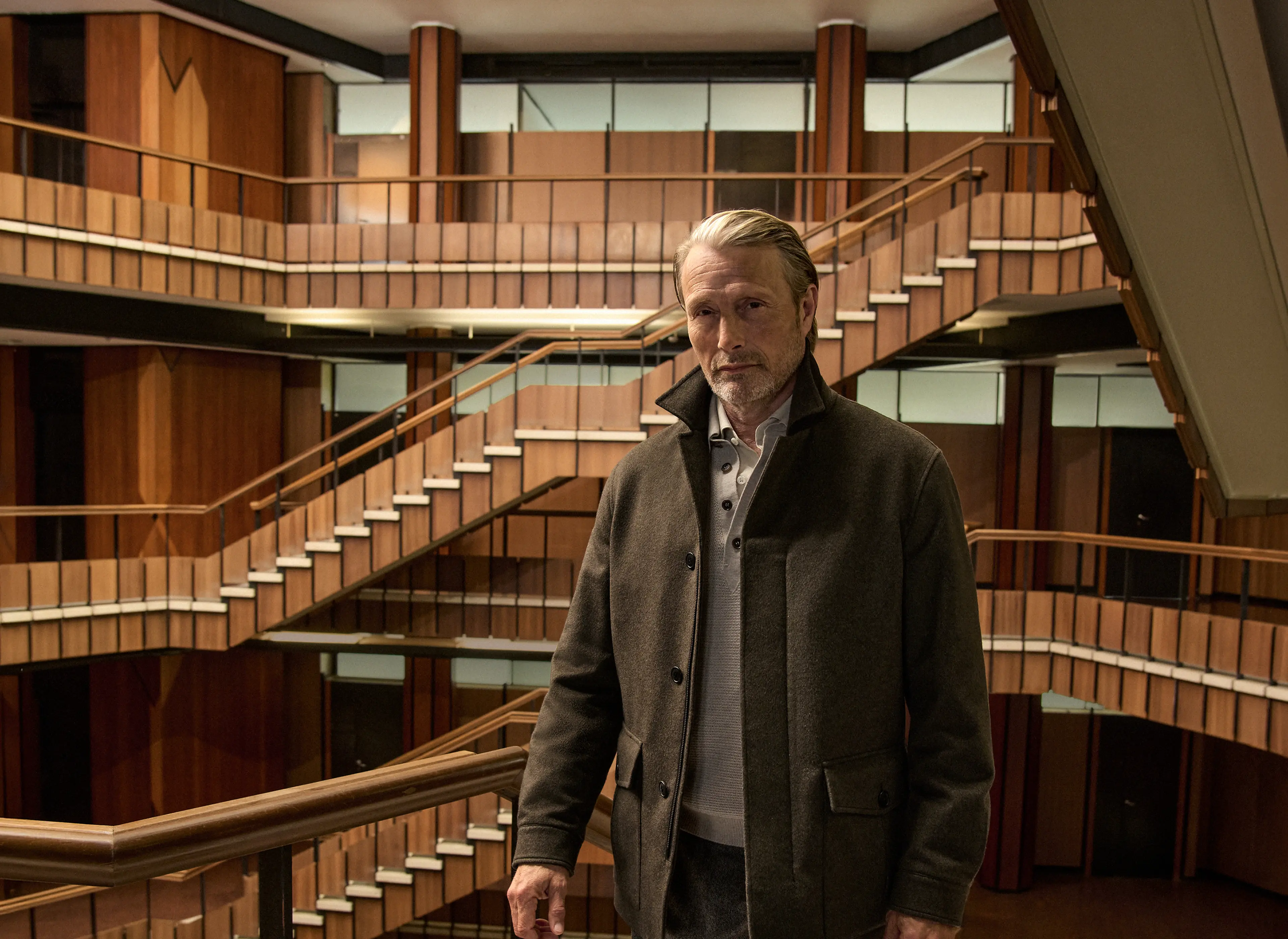 Mads Mikkelsen 身穿深橄榄色军装夹克，内搭针织衫，下着剪裁考究的裤子，脚蹬 Triple Stitch 鞋履，立于木质镶板中庭的楼梯之上，造型出自 ZEGNA（杰尼亚）2026 秋冬系列。