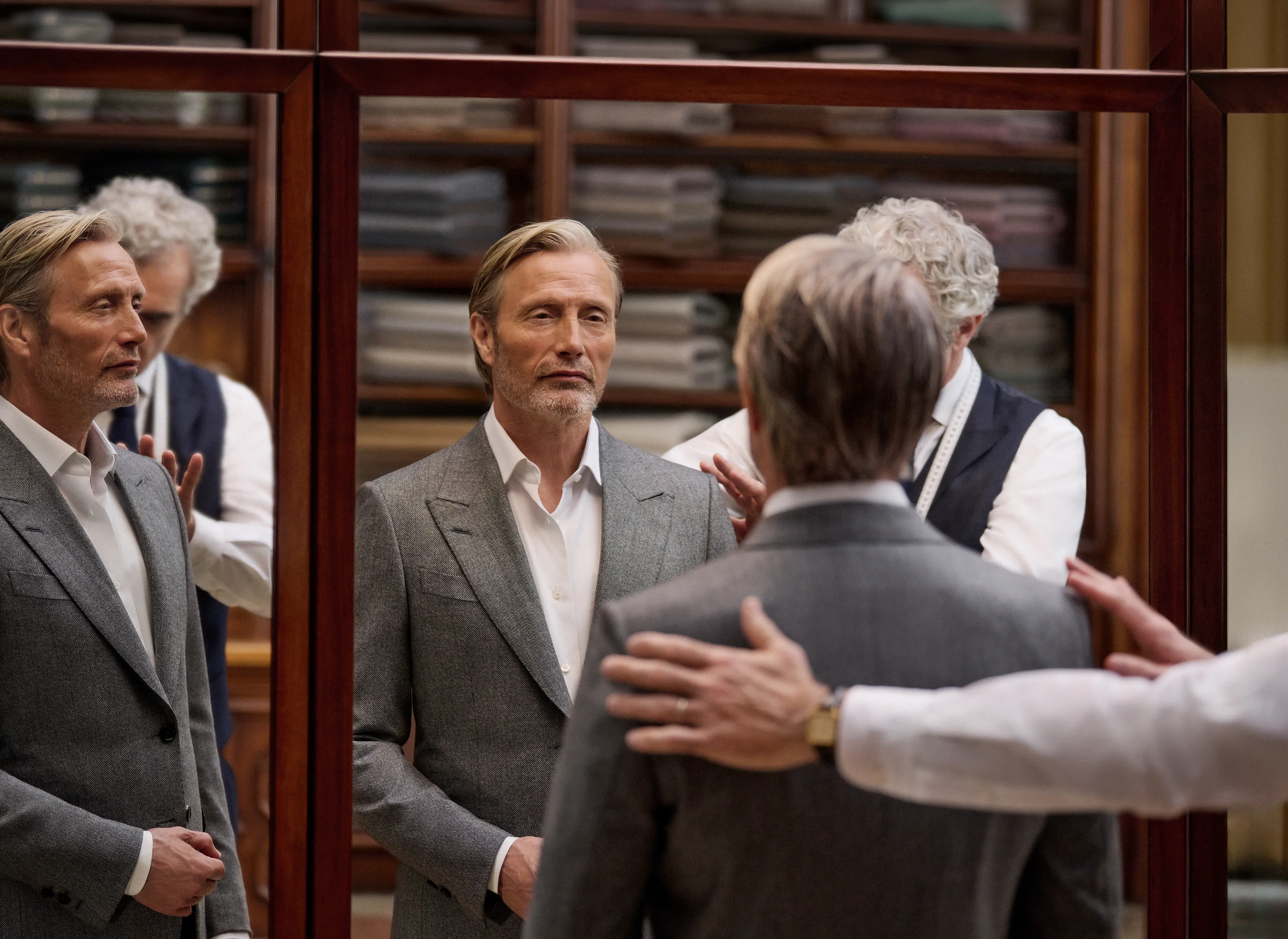 Mads Mikkelsen 身穿 ZEGNA 2026 秋冬系列灰色精裁西裝，在工作室內由裁縫師為其度身，倒映在試身室鏡中。