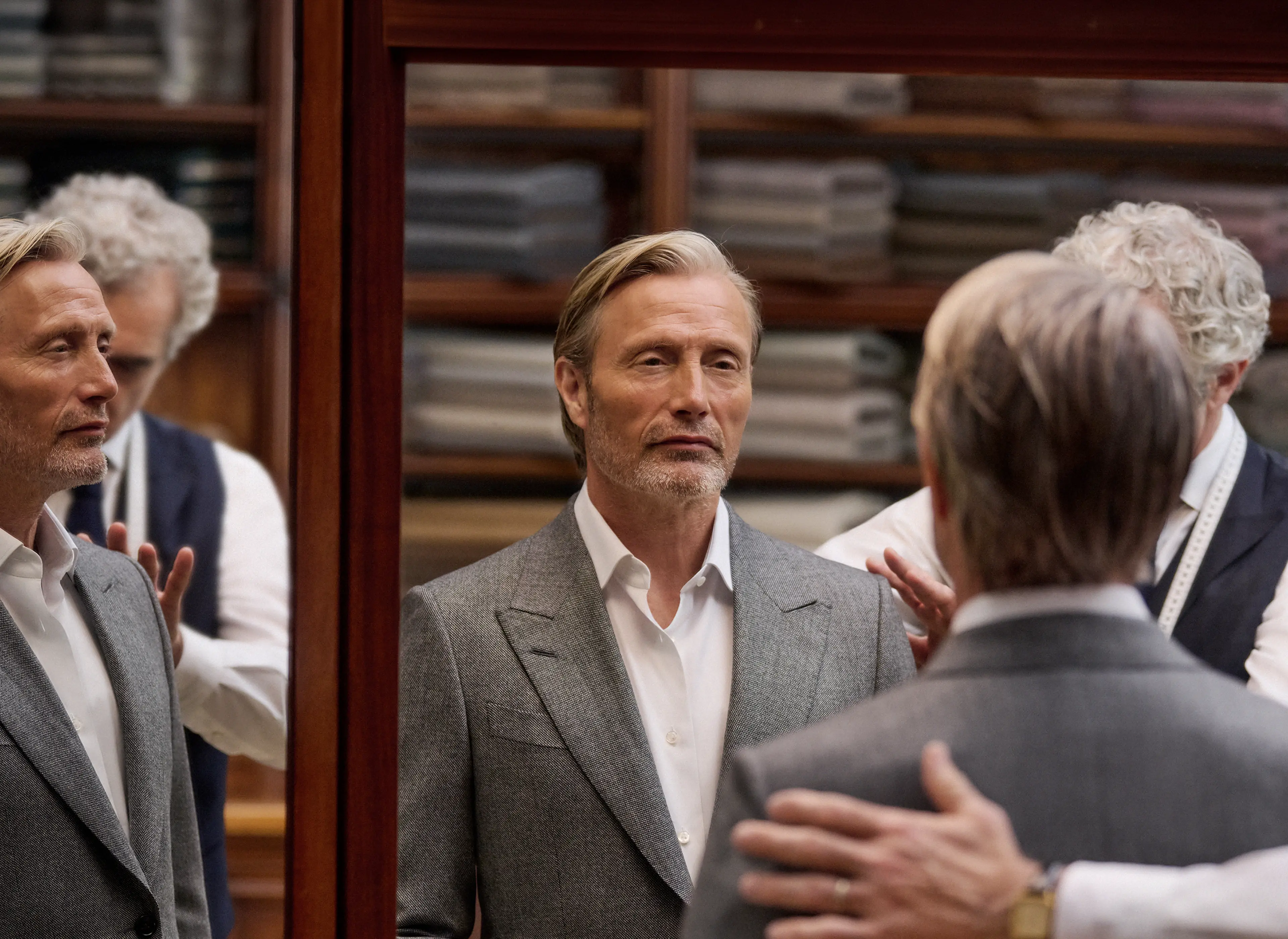 Mads Mikkelsen 身着灰色定制西装套装，于工坊内接受裁缝试装，身影映照在试衣间镜面之中，此造型出自 ZEGNA（杰尼亚）2026 秋冬系列。