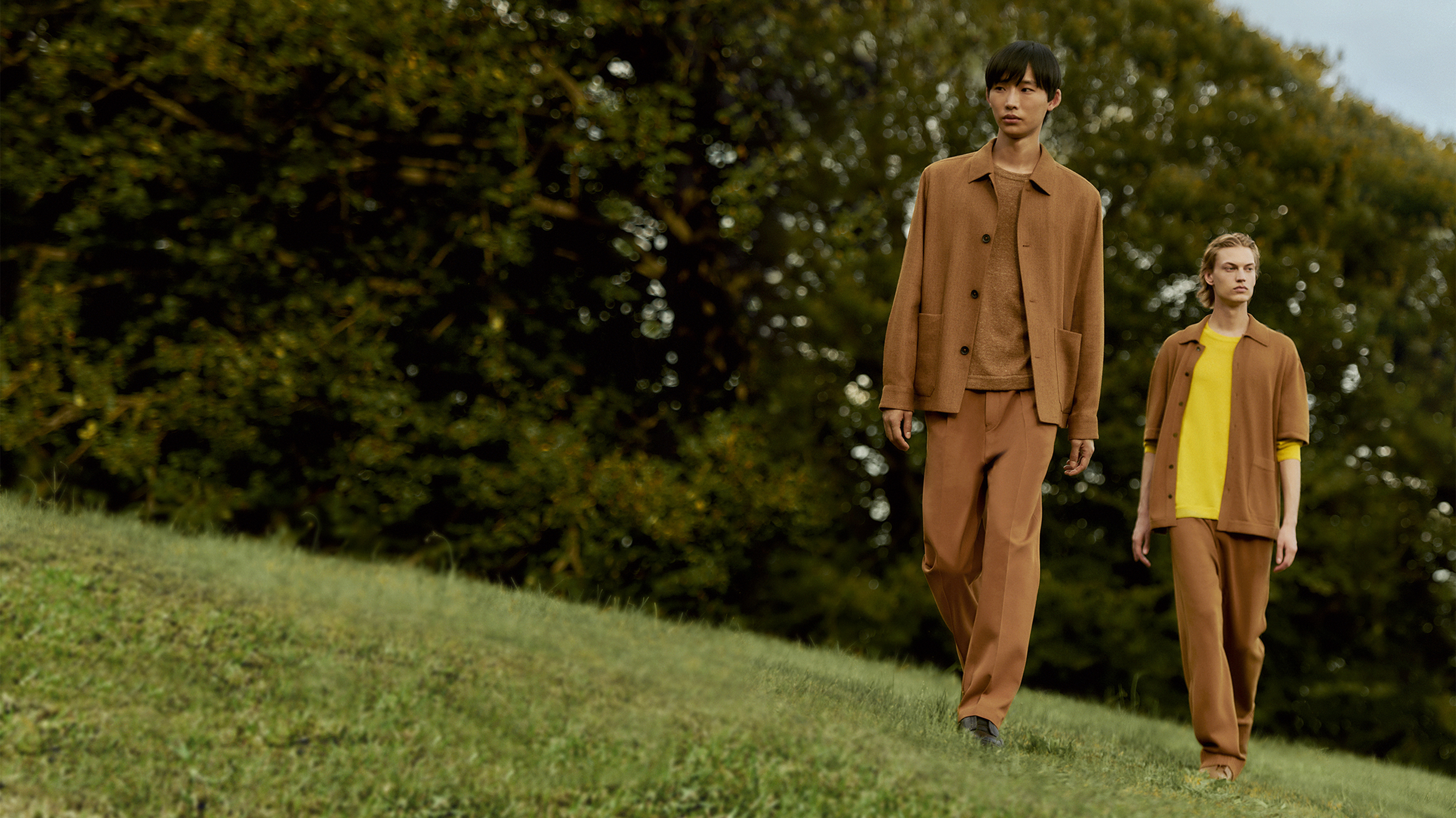 两位穿着 ZEGNA(杰尼亚)Oasi Cashmere 系列棕色 Overshirt 和长裤的男士在草地上行走。